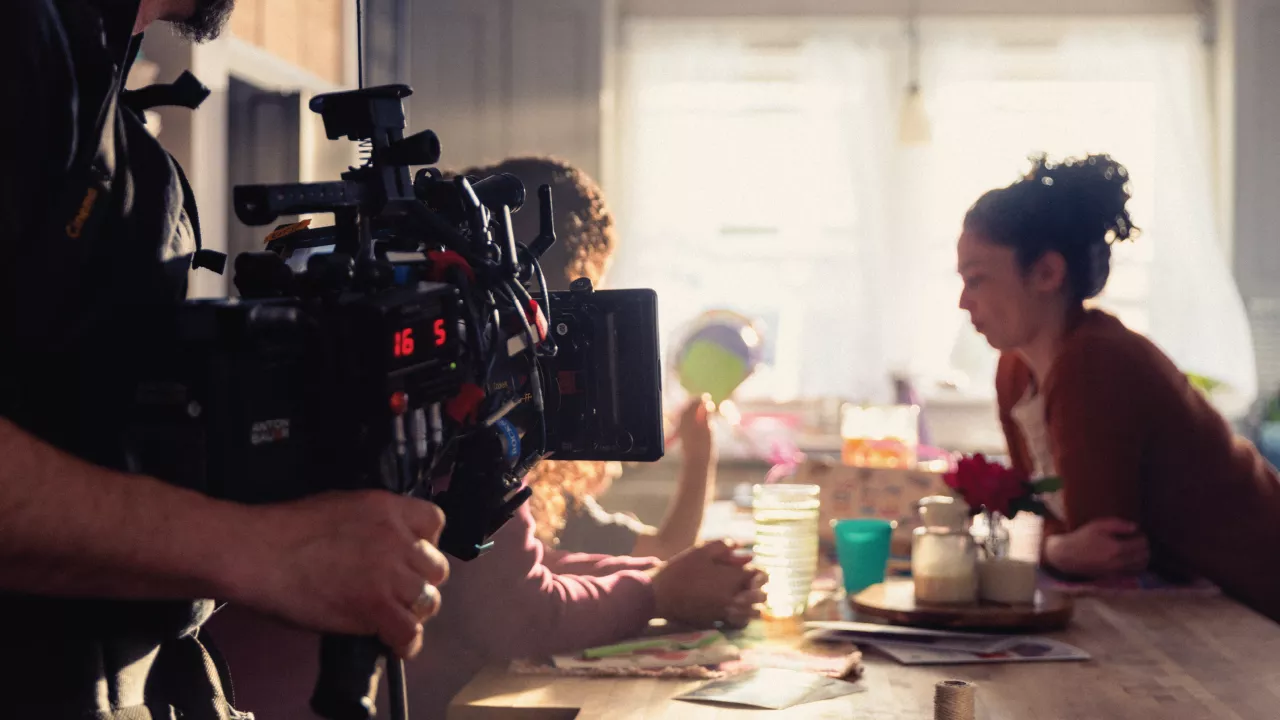 A camera operator films a kitchen scene while two people sit at a table in a softly lit room.