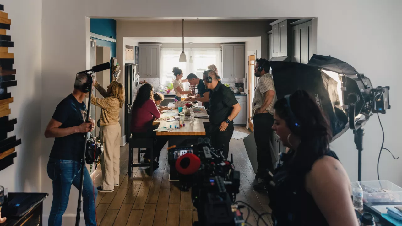 A wide view of a production crew filming a scene in a kitchen with various people involved in the shoot.