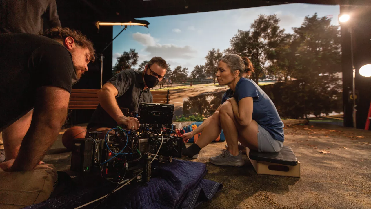 A director and crew examine a camera setup while an actor prepares in front of a green screen with a basketball court scene projected behind.