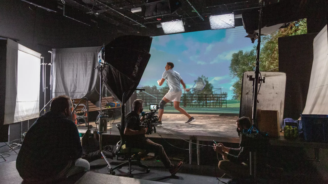 A tennis player prepares to swing during a virtual production scene, with a digitally projected background of a tennis court.