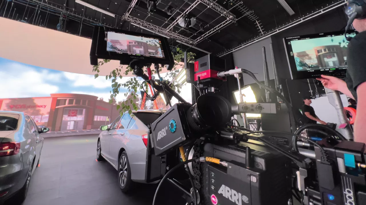 A camera captures a driving scene with multiple vehicles, filmed inside a virtual production studio featuring a background projection of a CVS pharmacy.